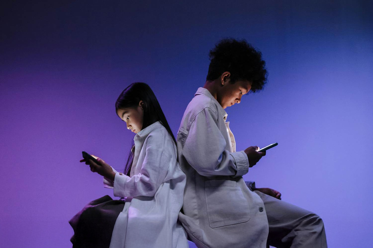 Dwoje dzieci siedzących i opartych plecami do siebie przeglądające smartfony na fioletowym tle.